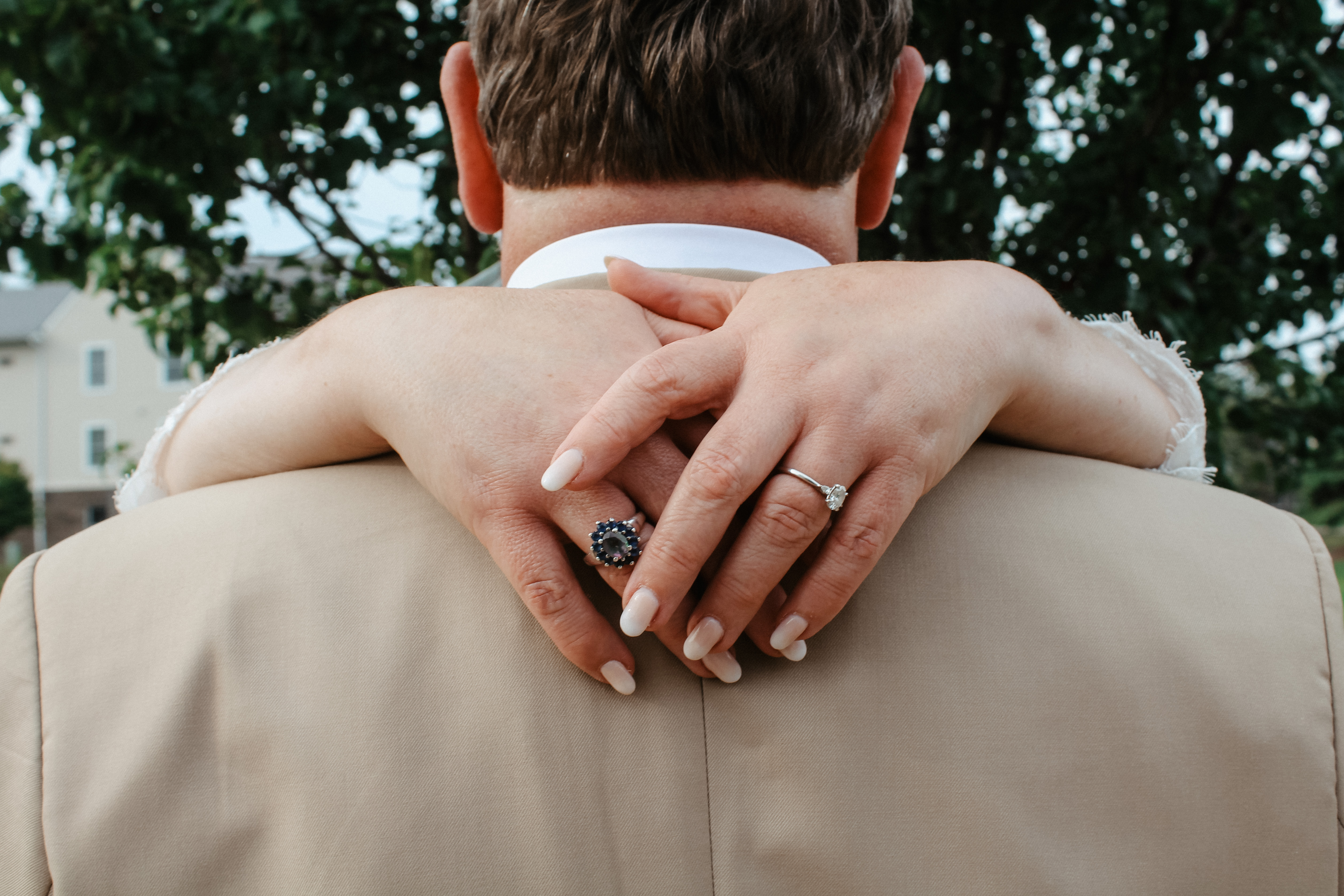 Closeup of bride’s hands around groom’s shoulders, showing engagement and wedding rings