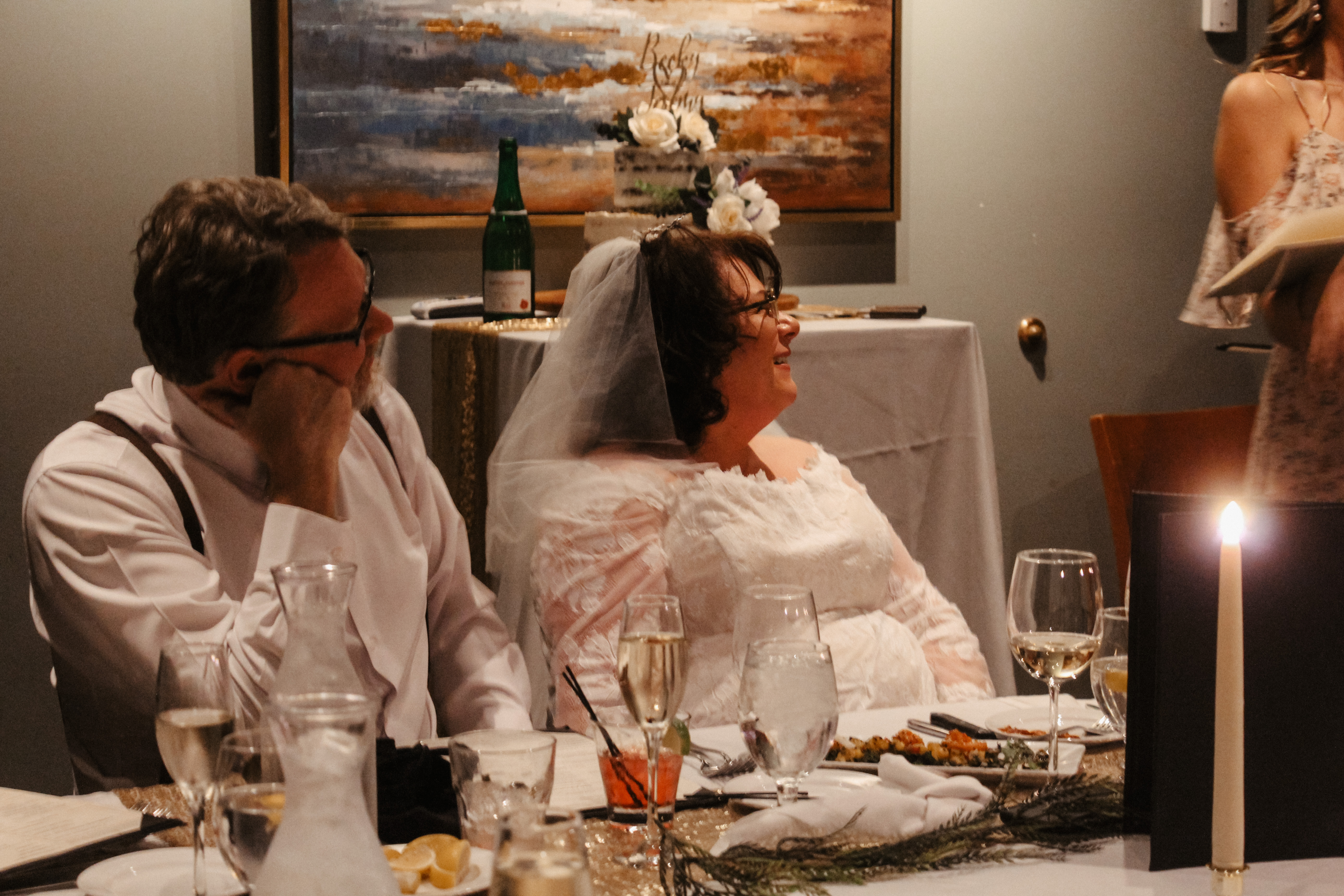 Bride and groom at wedding reception, laughing at table, wedding cake in background
